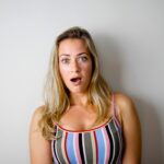 A young woman with a shocked expression stands against a plain white wall.