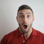 Portrait of a shocked man in a red polo shirt with a white background, expressing surprise.
